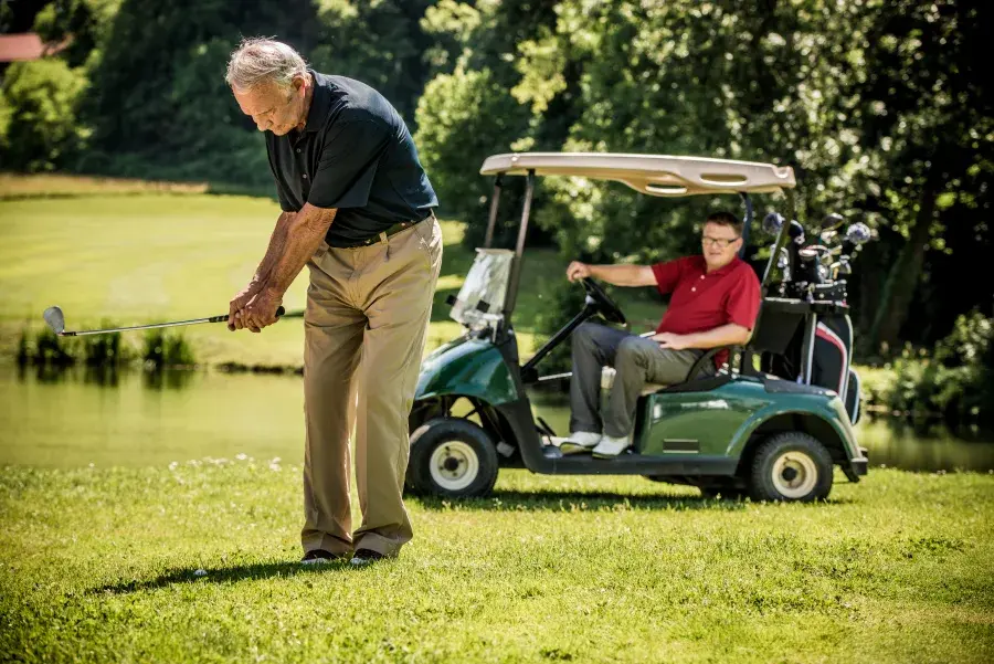 Two people golfing, one is hitting a ball while the other waits in the golf cart behind him